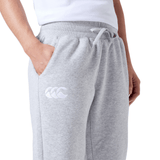 A person is wearing a pair of Women's Leisure Sweatpant by Canterbury, made from a cozy cotton blend fleece. The sweatpants feature a white drawstring and have the Canterbury logo on the pocket. Only their lower torso and hand in the pocket are visible.