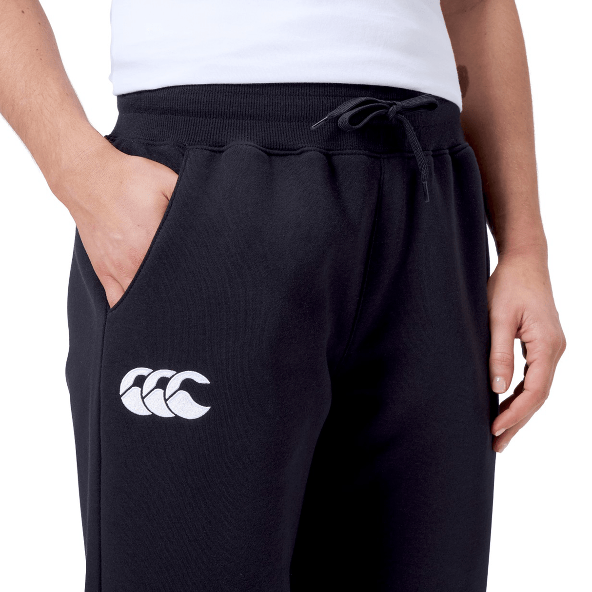A person wears the Women's Leisure Sweatpant by Canterbury, made from a cozy cotton blend fleece in black, featuring a white Canterbury logo on the left thigh and has a hand in one pocket.