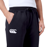 A person wears the Women's Leisure Sweatpant by Canterbury, made from a cozy cotton blend fleece in black, featuring a white Canterbury logo on the left thigh and has a hand in one pocket.