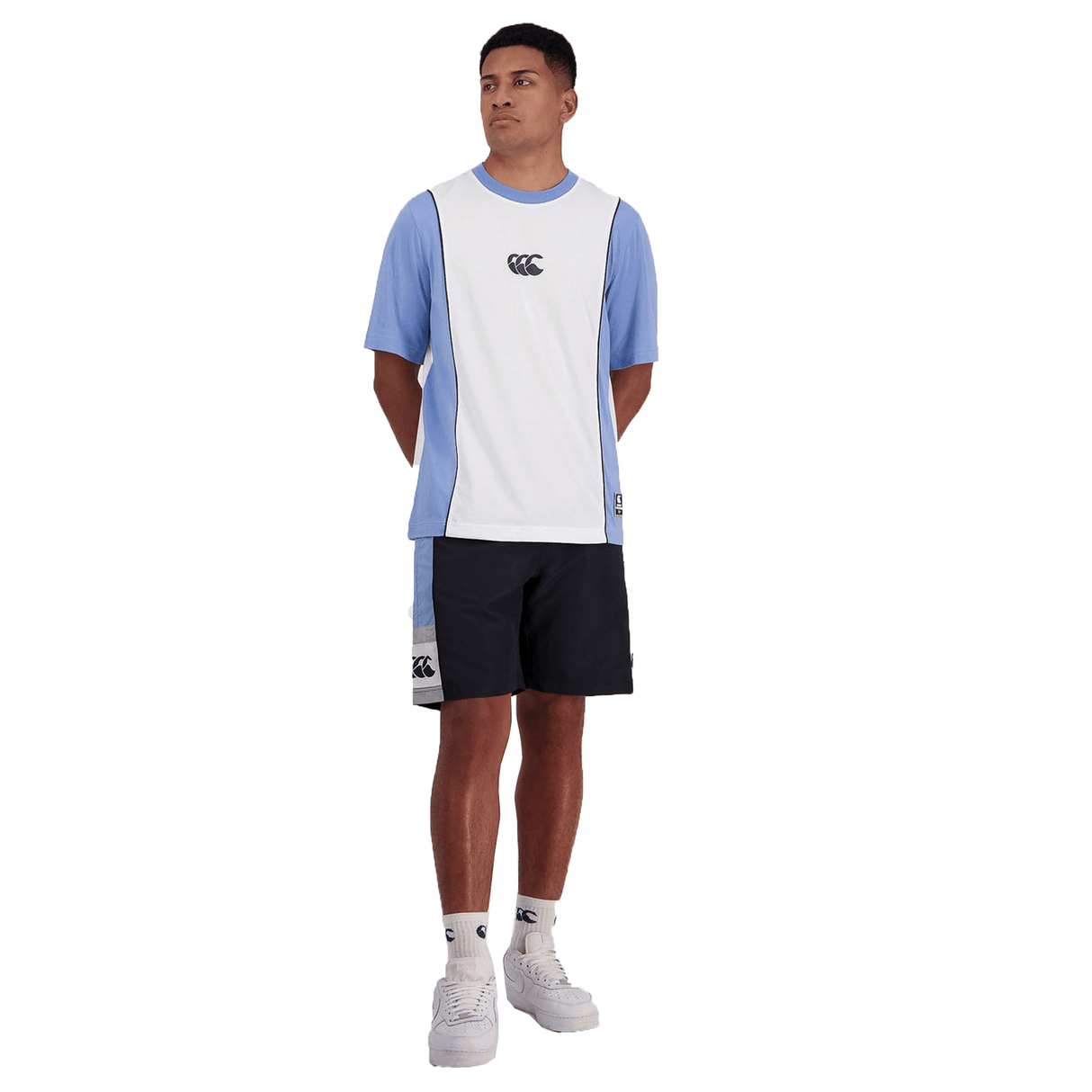 Man standing in a studio wearing a two-tone blue and white t-shirt, Canterbury Legends Panel Shorts with an elastic waistband, and white sneakers.