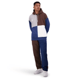 A person stands wearing a color-blocked hoodie in brown, blue, and white, paired with the Canterbury Harlequin Block Stadium Trackpant and white sneakers. They are looking upwards with hands in their pockets.