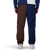 A person wearing Canterbury Harlequin Block Stadium Trackpants, featuring one leg in brown and the other in navy blue, facing away from the camera. The person is also sporting white sneakers and a color-blocked jacket.