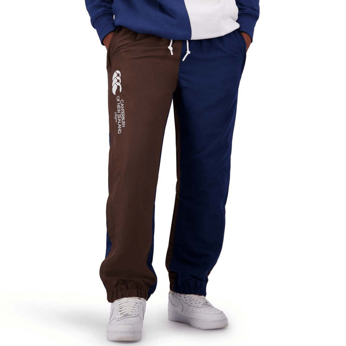 Person wearing Canterbury Harlequin Block Stadium Trackpant in blue and brown with white sneakers, proudly displaying the CCC logo on the left leg.