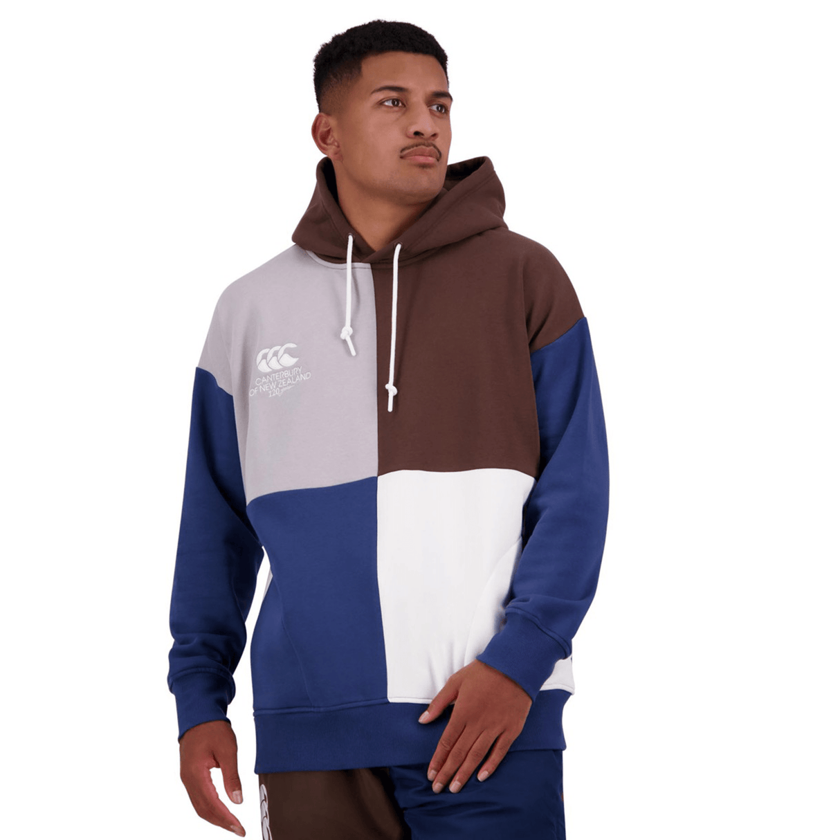A person is wearing a Canterbury Harlequin Block Pullover Hoodie with brown, gray, blue, and white sections and navy blue pants. The hoodie features a white Canterbury logo on the chest. The person is looking to the side.