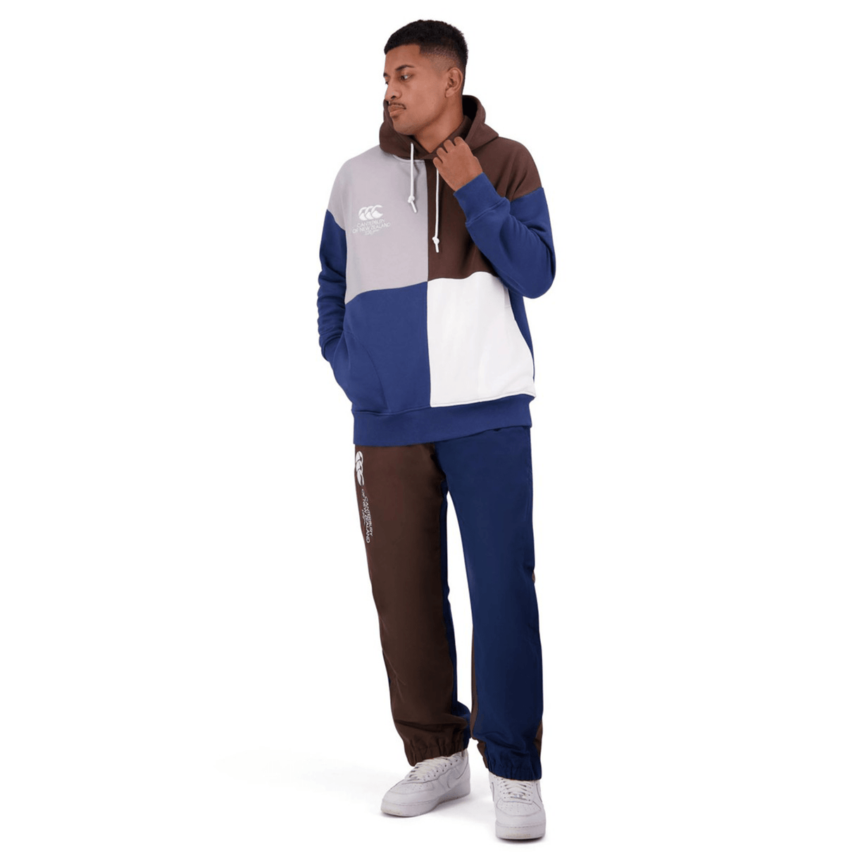 A man wearing a Canterbury Harlequin Block Pullover Hoodie featuring blue, gray, brown, and white blocks, paired with matching pants and white sneakers. He faces slightly to the left with one hand in his pocket.