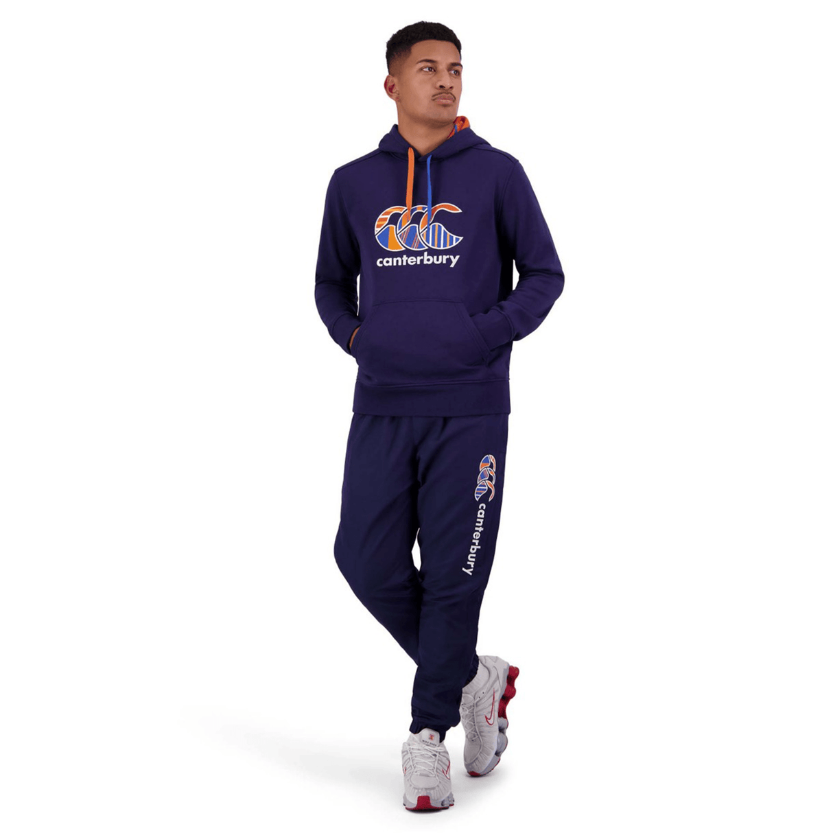 A person is standing against a white background, dressed in a navy blue Canterbury tracksuit and Canterbury Uglies Stadium Pants featuring a tapered leg, paired with sports shoes.
