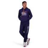 A person is standing against a white background, dressed in a navy blue Canterbury tracksuit and Canterbury Uglies Stadium Pants featuring a tapered leg, paired with sports shoes.