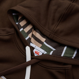 The Canterbury Uglies Hoodie features a brown color with a white drawstring, and a hood with a green, beige, and brown striped inner lining that showcases the stylized CCC logo next to the garment tag.