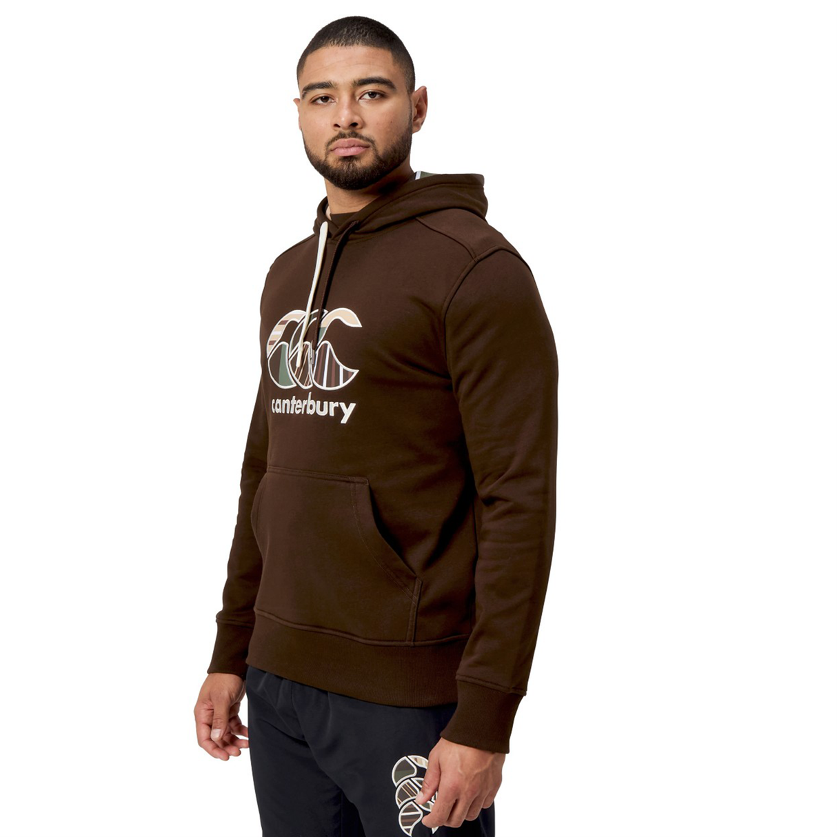 A person wearing a Canterbury Uglies Hoodie, featuring a brown color and a stylized CCC logo, stands slightly turned with hands by their sides and neutral expression.