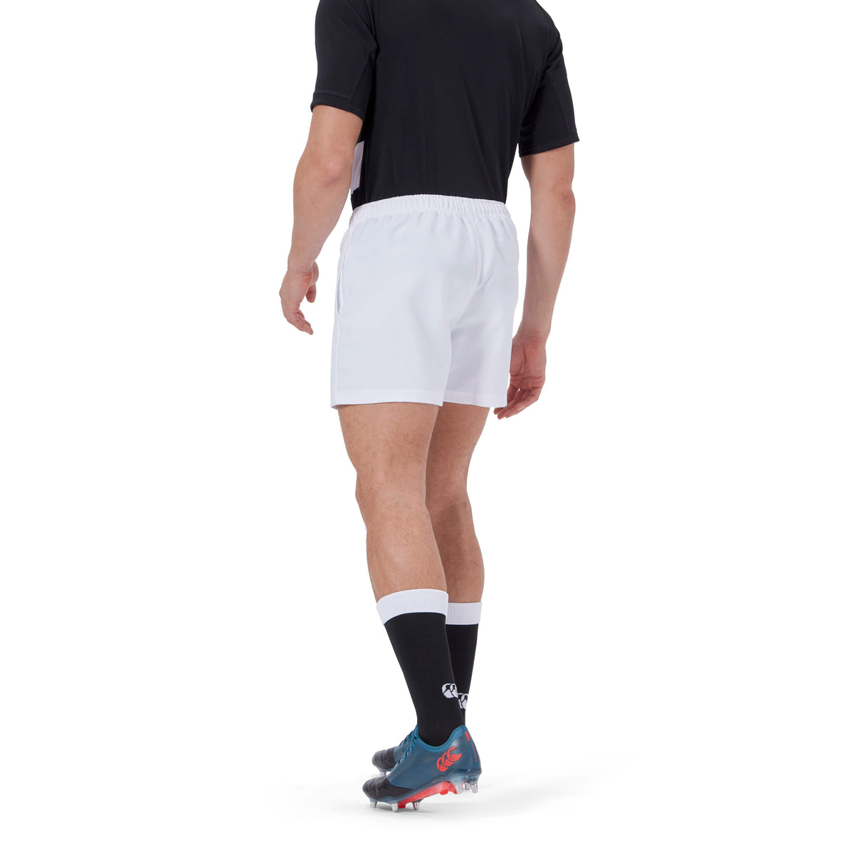 A person is standing with their back to the camera, wearing a black shirt, Canterbury Player's Drill Short in white, black socks, and cleated shoes crafted by Canterbury.