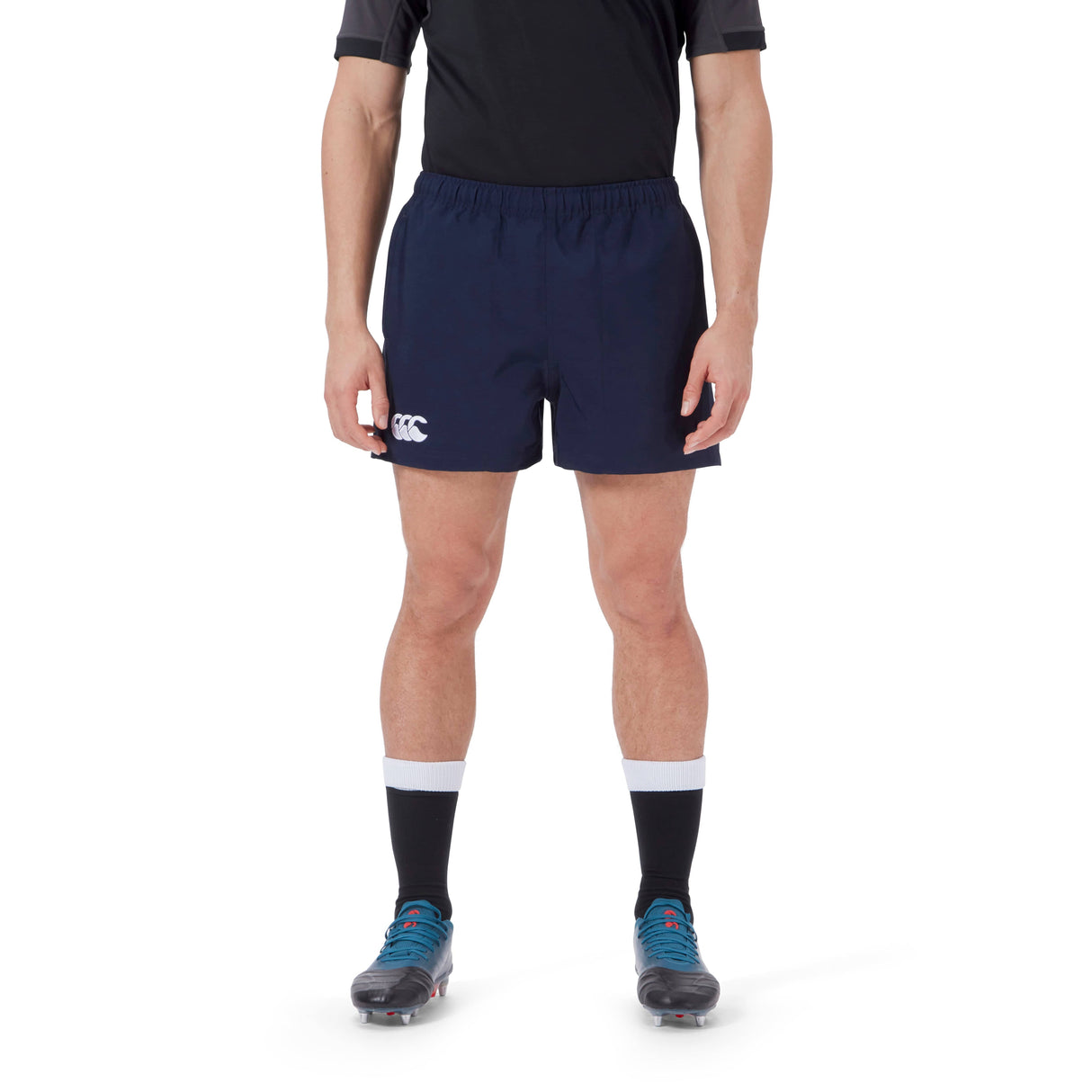 A person wearing a black shirt, navy blue Canterbury Player's Drill Short with the "CCC" logo, black socks with white bands, and blue cleats stands against a white background.