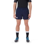 A person wearing a black shirt, navy blue Canterbury Player's Drill Short with the "CCC" logo, black socks with white bands, and blue cleats stands against a white background.