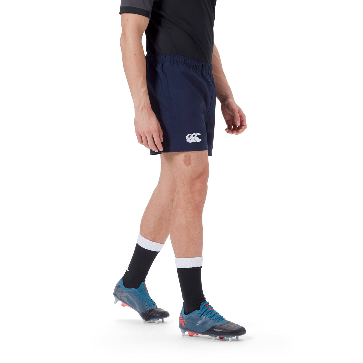 A person wearing a black shirt, Canterbury Player's Drill Short in navy blue made from recycled fabric, black socks with white bands, and blue athletic shoes stands on a white background.