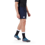 A person wearing a black shirt, Canterbury Player's Drill Short in navy blue made from recycled fabric, black socks with white bands, and blue athletic shoes stands on a white background.