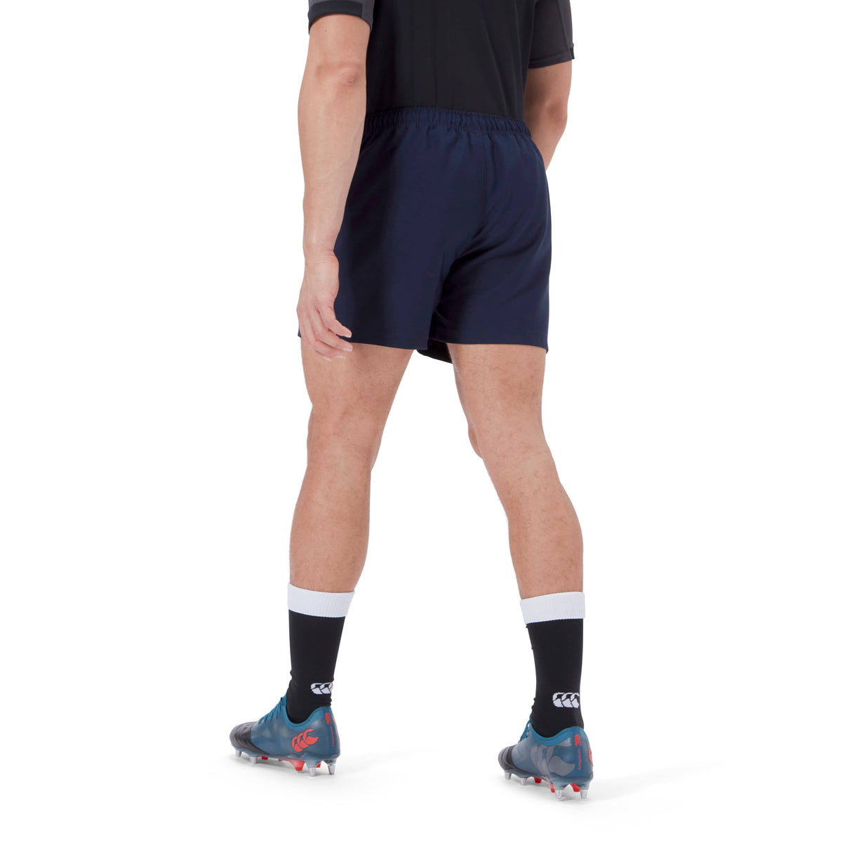 A person wearing blue Canterbury Player's Drill Shorts, blue and white soccer socks, black soccer shoes, and a black top stands with their back to the camera.