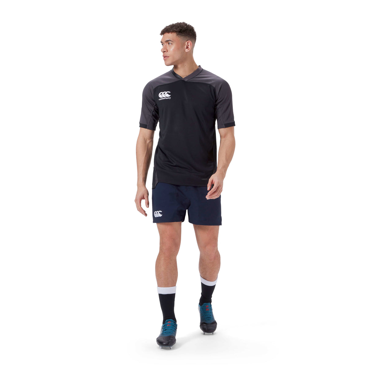 A man wearing a black Canterbury sports shirt, Canterbury Player's Drill Short, black socks, and blue shoes stands on a white background.