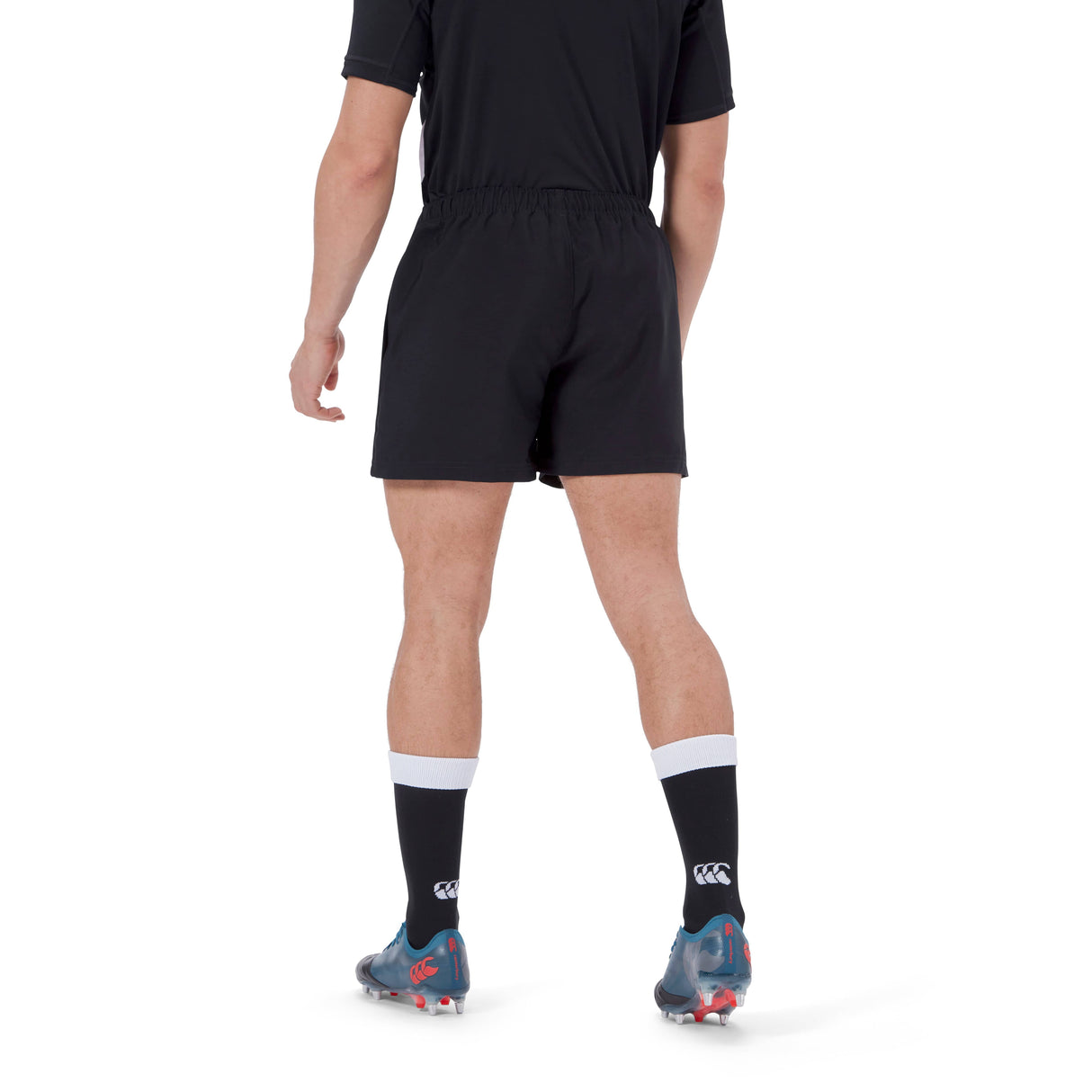 A person stands facing away, wearing black Canterbury Player's Drill Short, a black shirt, black socks with white tops, and gray sports shoes on a white background.