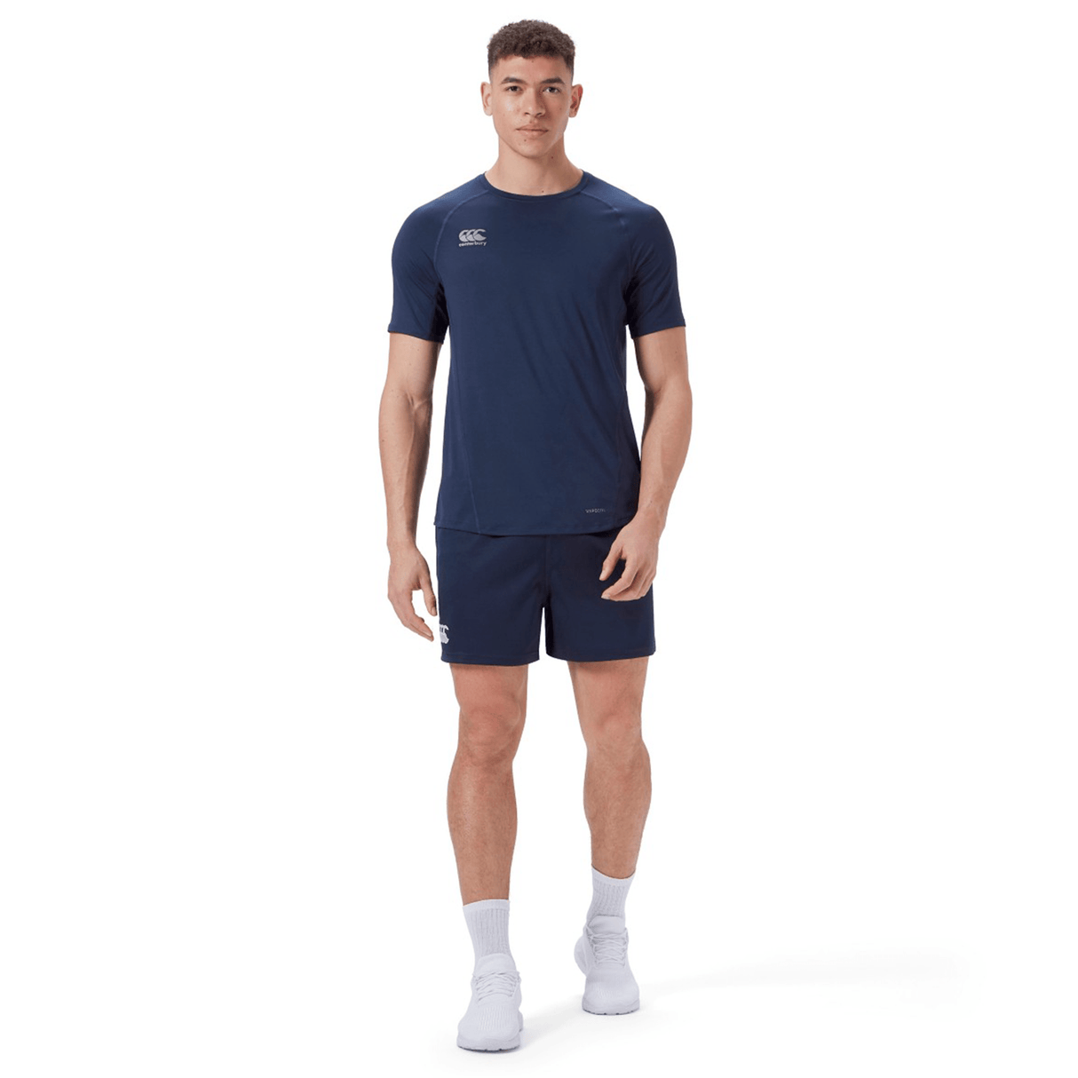 A man stands facing forward against a plain white background, wearing Canterbury's Professional Short 2.0 in navy and an athletic t-shirt with an adjustable fit, paired with white socks and sneakers.