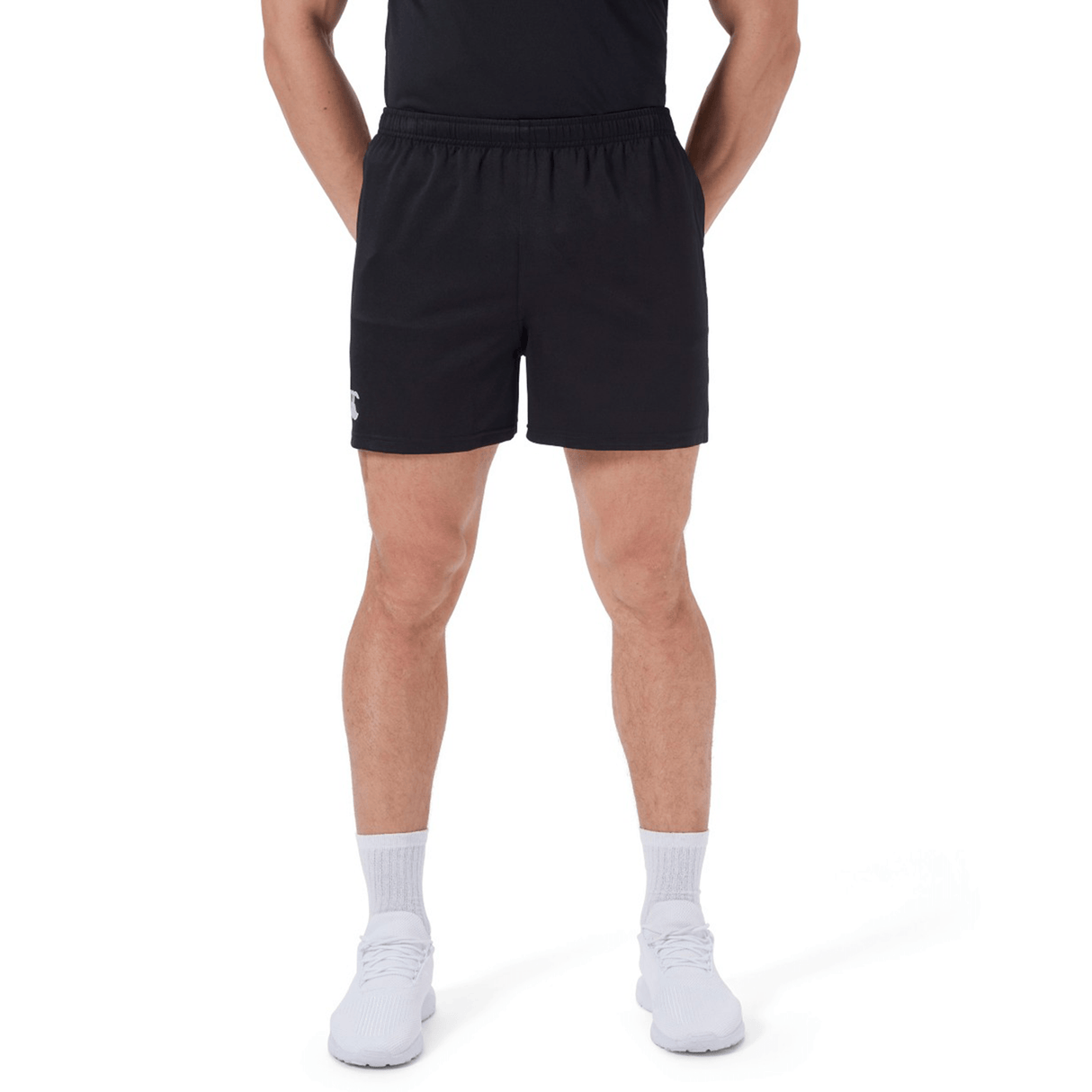 A person wearing black Canterbury Professional Short 2.0, a black shirt, white socks, and white athletic shoes, standing with hands behind their back.