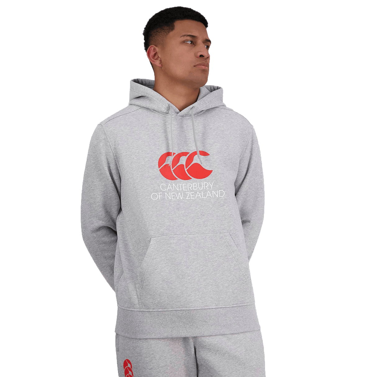 A person wearing a light grey Canterbury of New Zealand Hoodie by Canterbury with a red "Canterbury" logo and matching grey sweatpants stands with their hands behind their back, looking to the side.