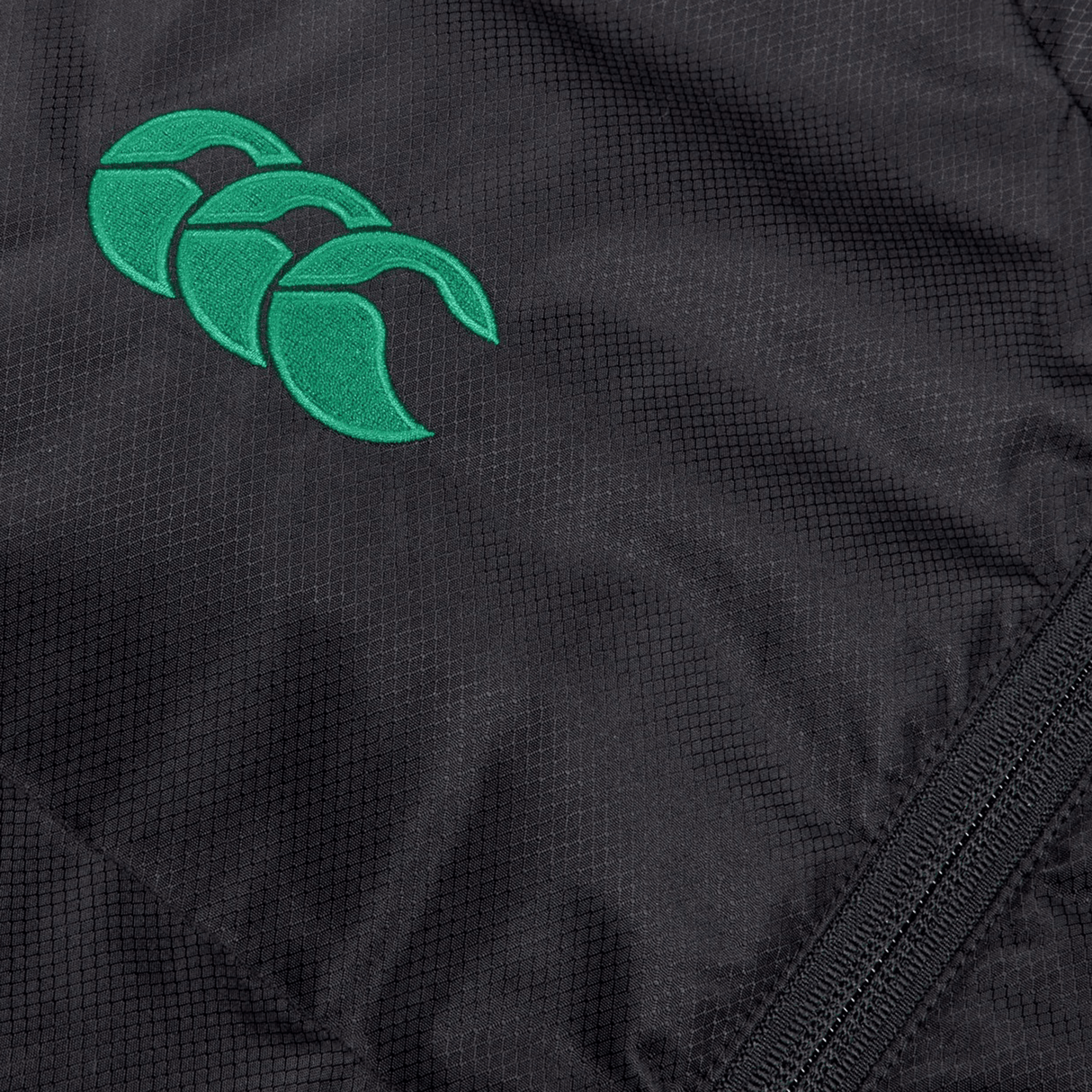Close-up of a black textured fabric with a green stitched logo featuring three interlocking crescents. A visible zipper lies along the right edge of the Ireland 24 Padded Gilet by Canterbury, enhanced by Vaposhield coating for added durability.