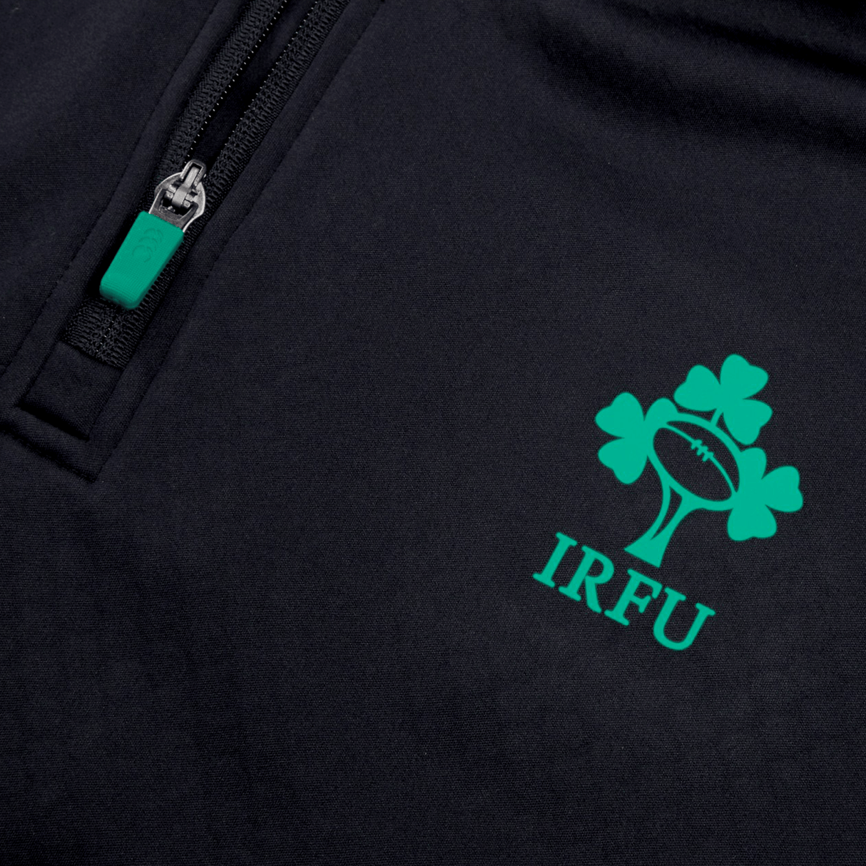 The Ireland 1/4 Zip Training Top 24 by Canterbury, featuring a dark fabric adorned with the IRFU logo—which includes a rugby ball and clover—along with a zipper that has a green pull tab, is perfect for enthusiasts.
