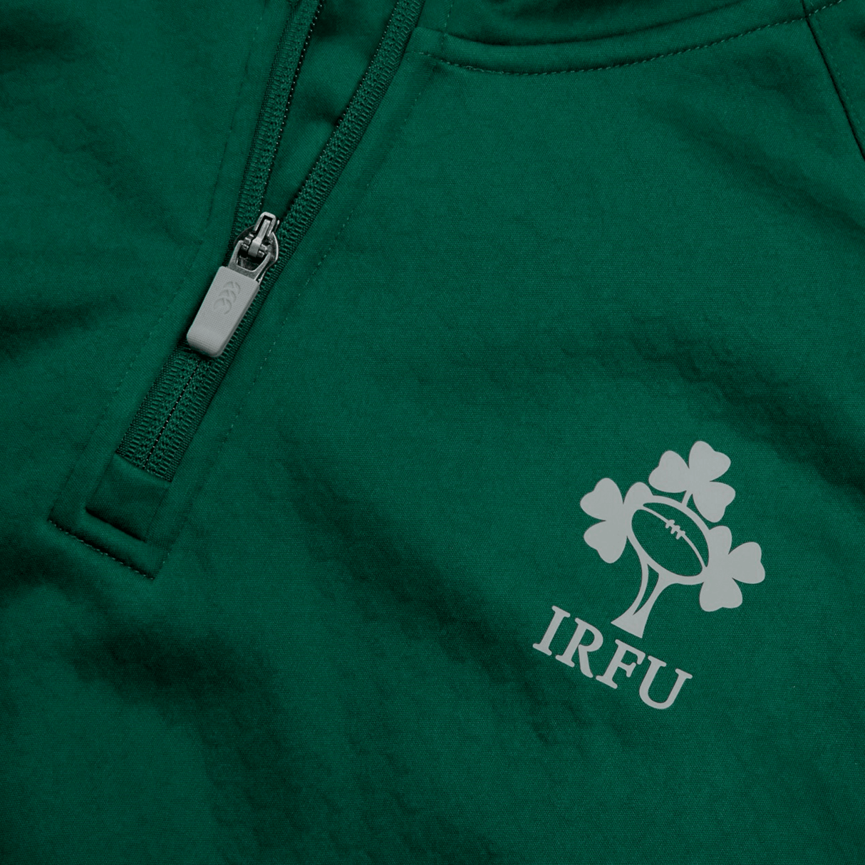 Close-up of the Ireland 1/4 Zip Training Top 24 by Canterbury in green, showcasing the IRFU logo featuring a rugby ball, shamrock, and the initials "IRFU," along with a zipper detail.