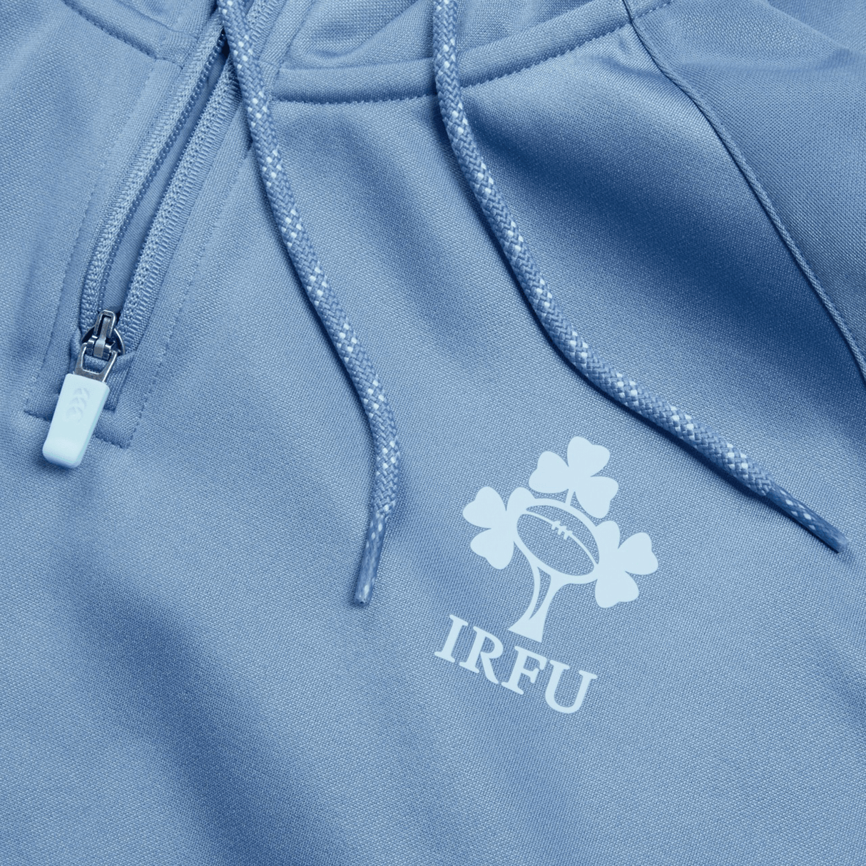 Close-up of a light blue Ireland Training Hoodie 24 by Canterbury, featuring a zipper and drawstrings. This durable polyester hoody showcases the IRFU logo with a rugby ball and a shamrock, making it perfect for showing your support while staying comfortable.