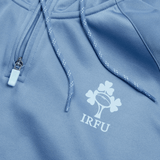 Close-up of a light blue Ireland Training Hoodie 24 by Canterbury, featuring a zipper and drawstrings. This durable polyester hoody showcases the IRFU logo with a rugby ball and a shamrock, making it perfect for showing your support while staying comfortable.