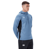 A man wearing a light blue Ireland Training Hoodie 24 by Canterbury and black pants stands against a plain white background.