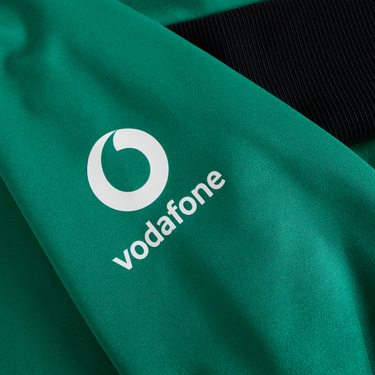 A green fabric from the Ireland 25/26 Training Hoodie by Canterbury featuring the white Vodafone logo, shown beside black ribbed material.