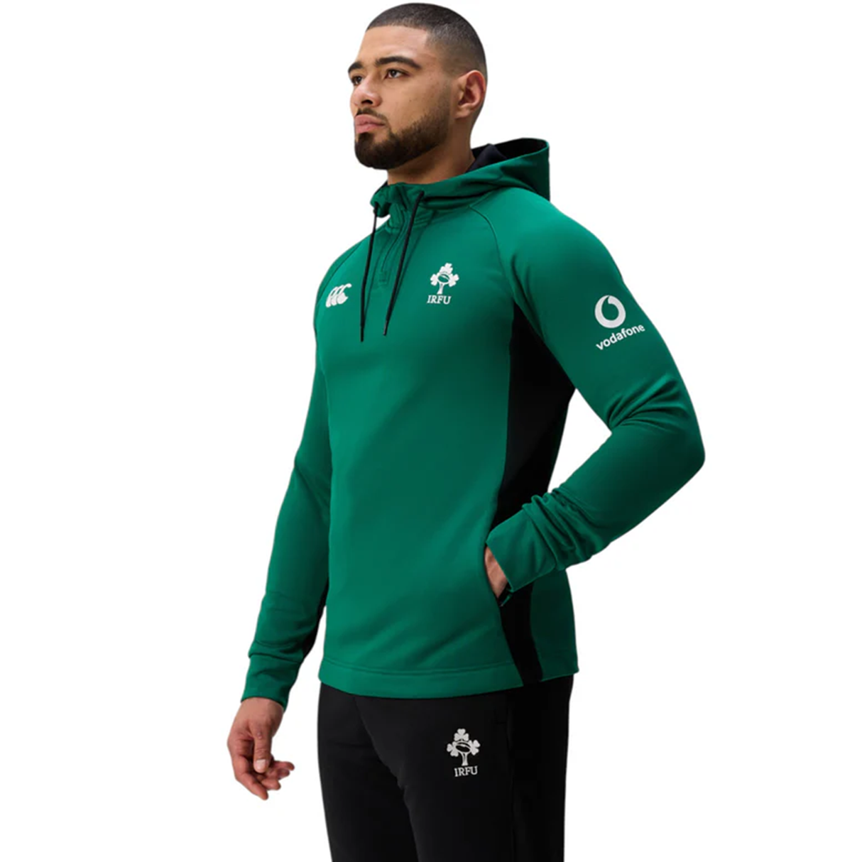 A man wears the Ireland 25/26 Training Hoodie by Canterbury in green with the IRFU crest and black pants with the IRFU logo, standing against a white background.