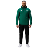 A man faces forward wearing the Ireland 25/26 Training Hoodie by Canterbury, featuring logos, black pants, and white athletic shoes, set against a plain white background.