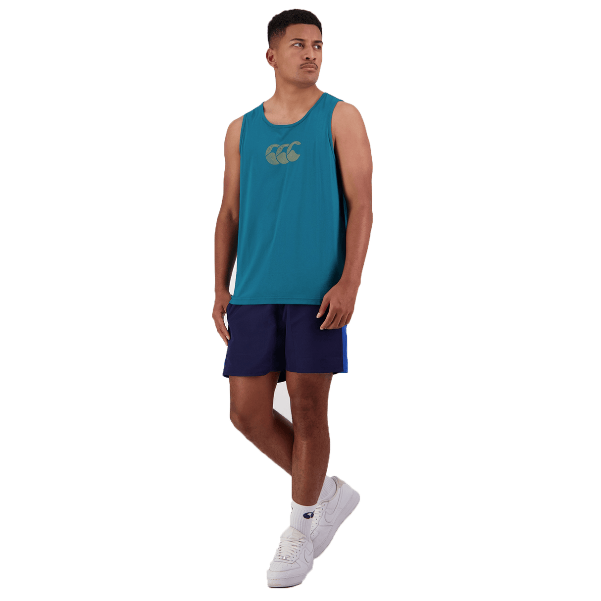A person is dressed in a Canterbury Radial CCC Logo Singlet in teal, navy shorts, and white sneakers, standing against a white background.