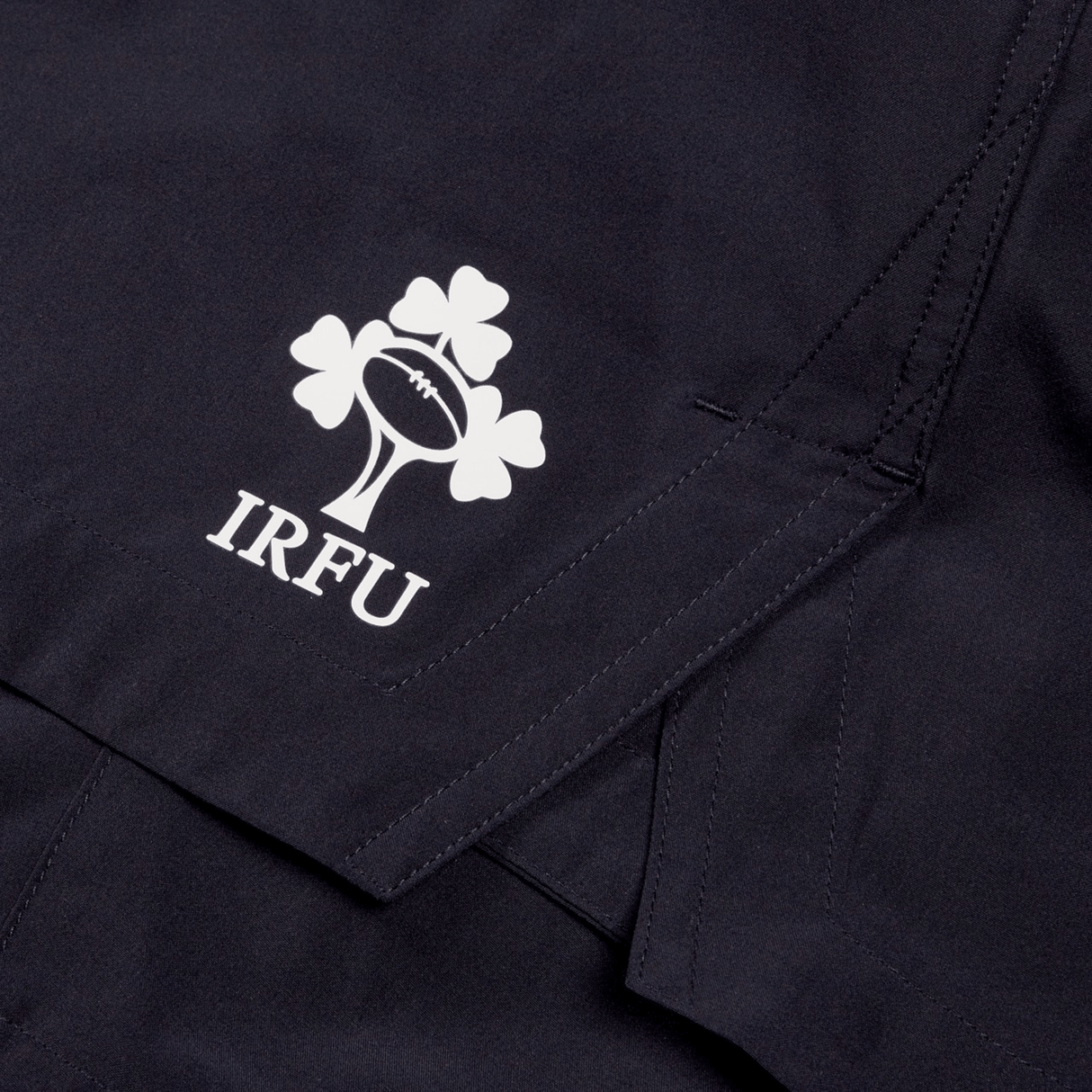 Close-up of a dark fabric with a white Irish Rugby Football Union (IRFU) logo, featuring a shamrock and rugby ball, printed on what appears to be the Ireland 24 Woven Gym Short by Canterbury.