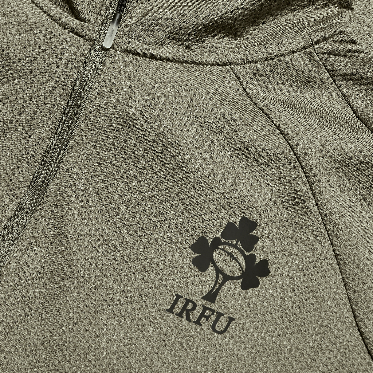 Close-up of the Ireland 25/26 Elite First Layer 1/4 Zip by Canterbury, featuring a textured gray fabric, zipper, and IRFU logo with rugby ball and shamrocks.