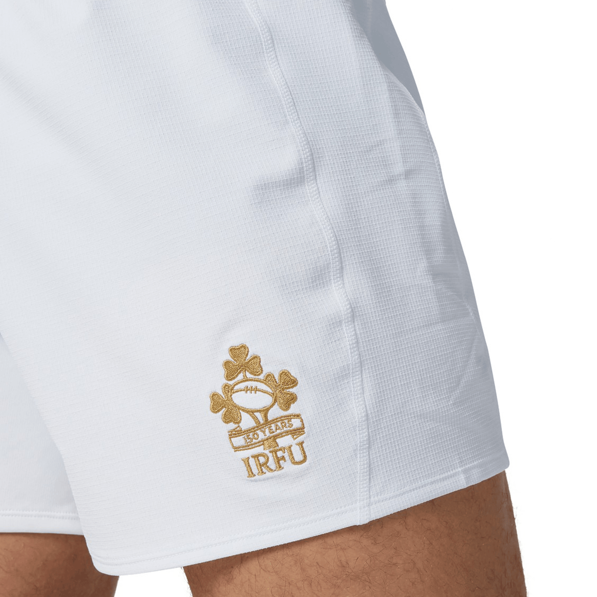 A close-up shows an individual sporting the Ireland 150th Anniversary Home Match Short by Canterbury, adorned with a gold embroidered emblem featuring a rugby ball, shamrocks, and the "150 Years IRFU" inscription. Ideal for celebrating Ireland's 150th Anniversary at an exhilarating home match.