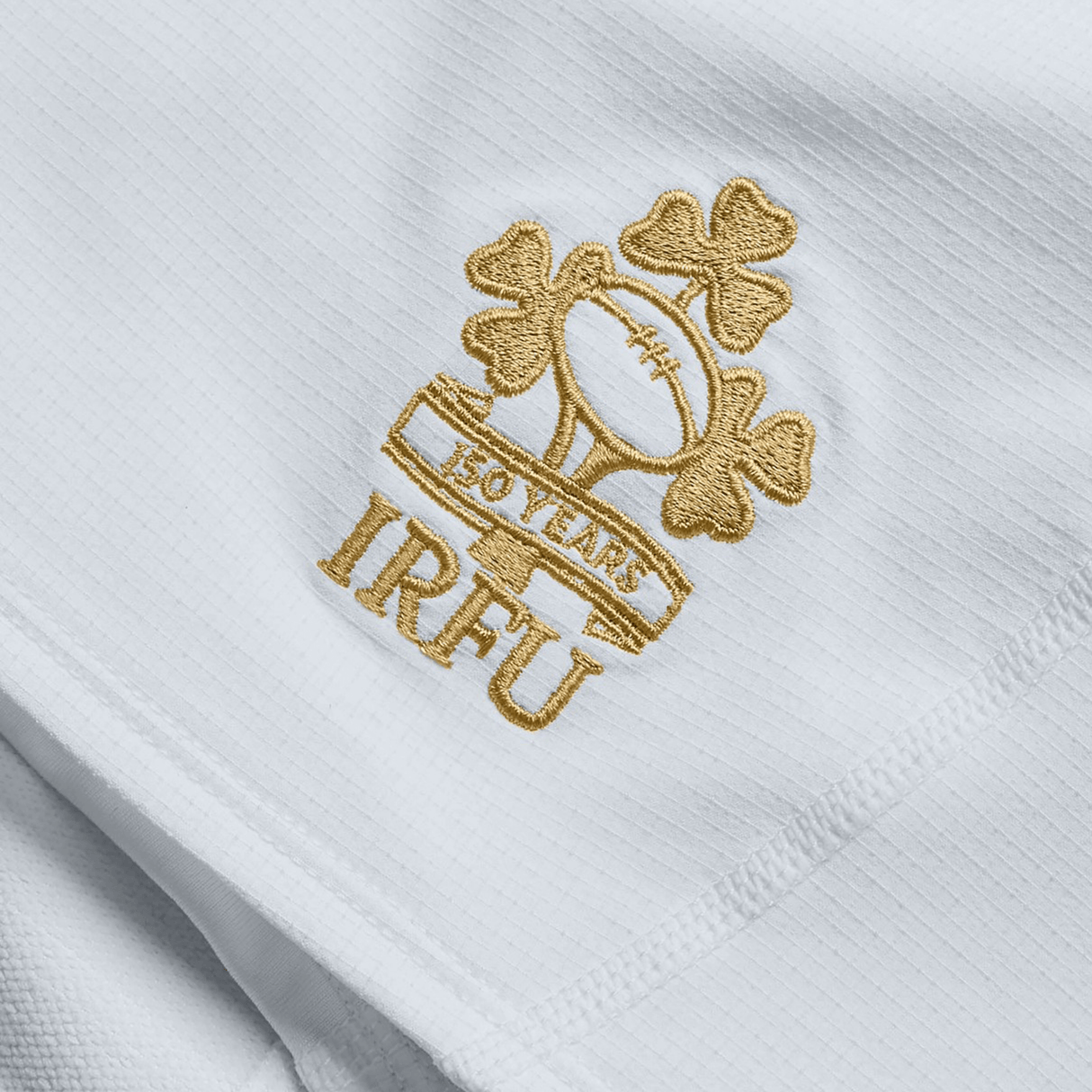 Close-up of a white fabric from Canterbury's Ireland 150th Anniversary Home Match Short, featuring an embroidered gold emblem with "IRFU," a rugby ball, shamrocks, and "150 Years" celebrating Ireland's milestone. Perfect for showcasing at any home match event.