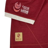 A close-up of the British & Irish Lions 2025 Players Authentic Jersey by Canterbury shows a red sports jersey featuring the Qatar Airways Lions Men's Series Australia 2025 logo on the sleeve and a lion emblem patch with text near the bottom, capturing the essence of the tour jersey.