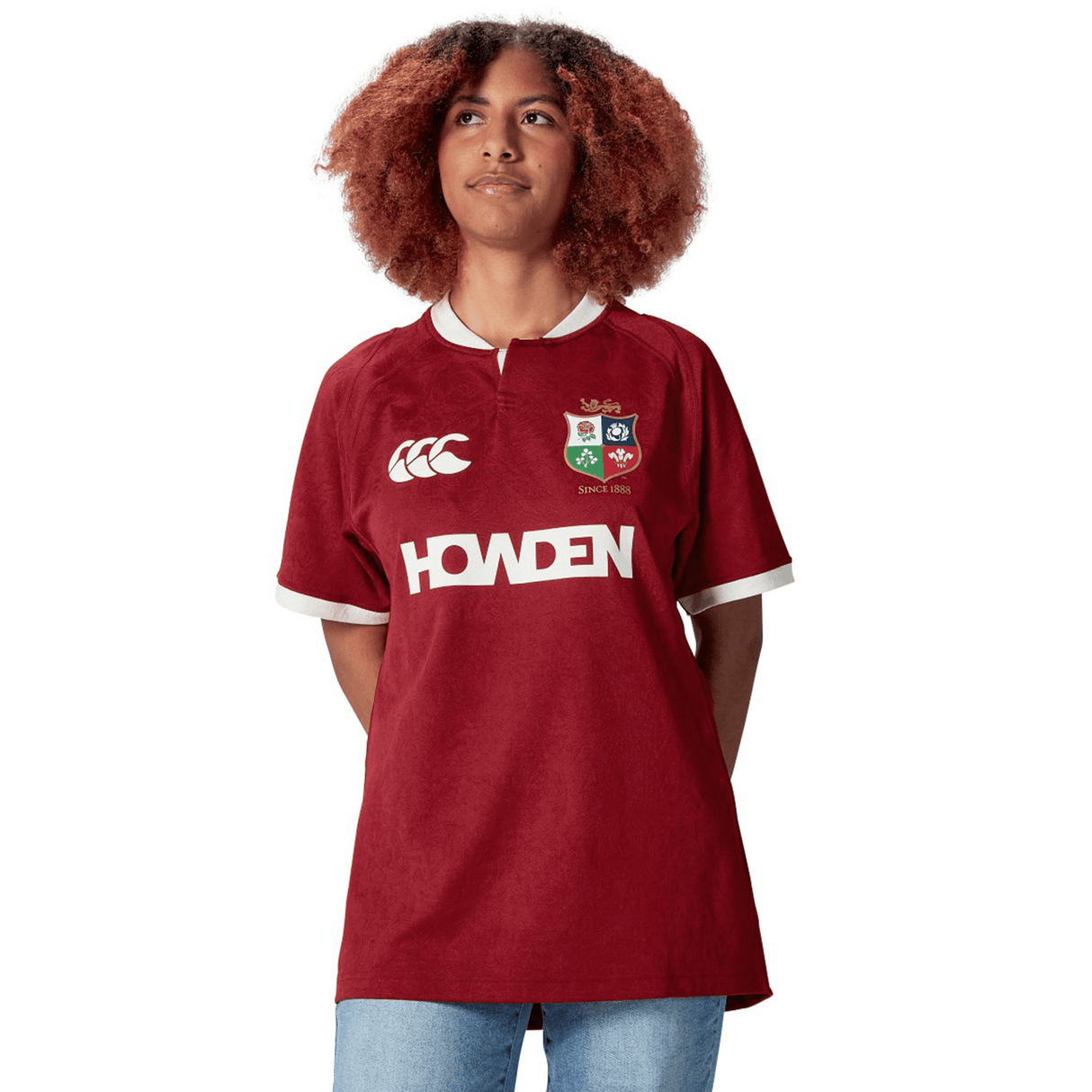A person with curly hair wears a red British & Irish Lions 2025 Players Jersey by Canterbury, featuring logos such as "Howden" and a crest, standing with their hands behind their back.