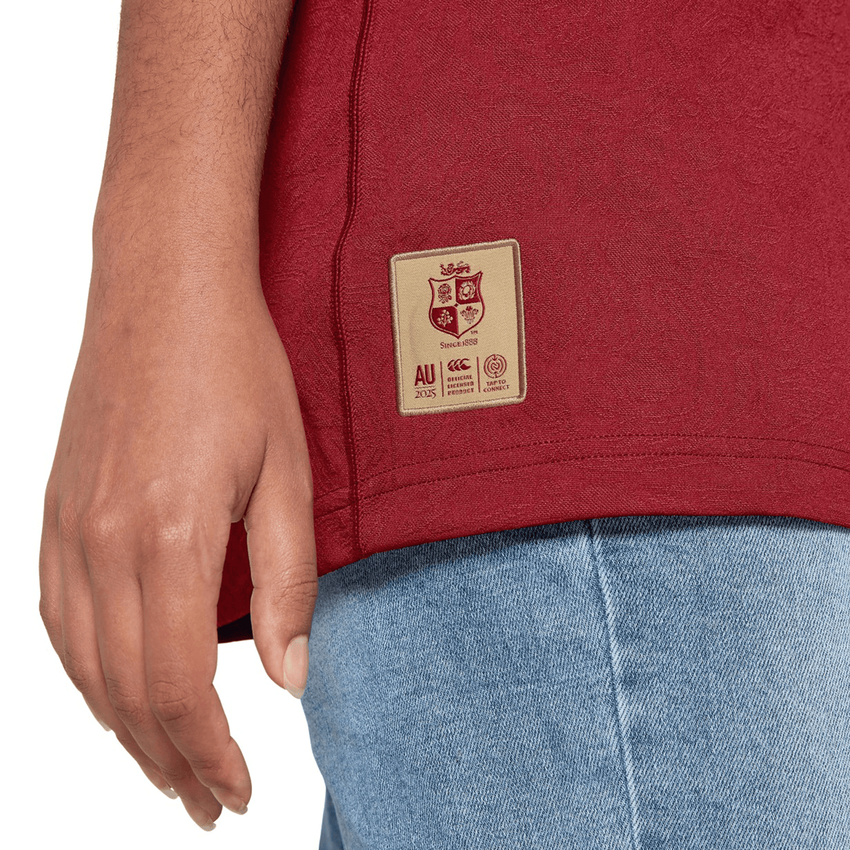 Close-up of a person wearing the British & Irish Lions 2025 Players Jersey by Canterbury, paired with blue jeans. Only the lower torso and hand are visible.