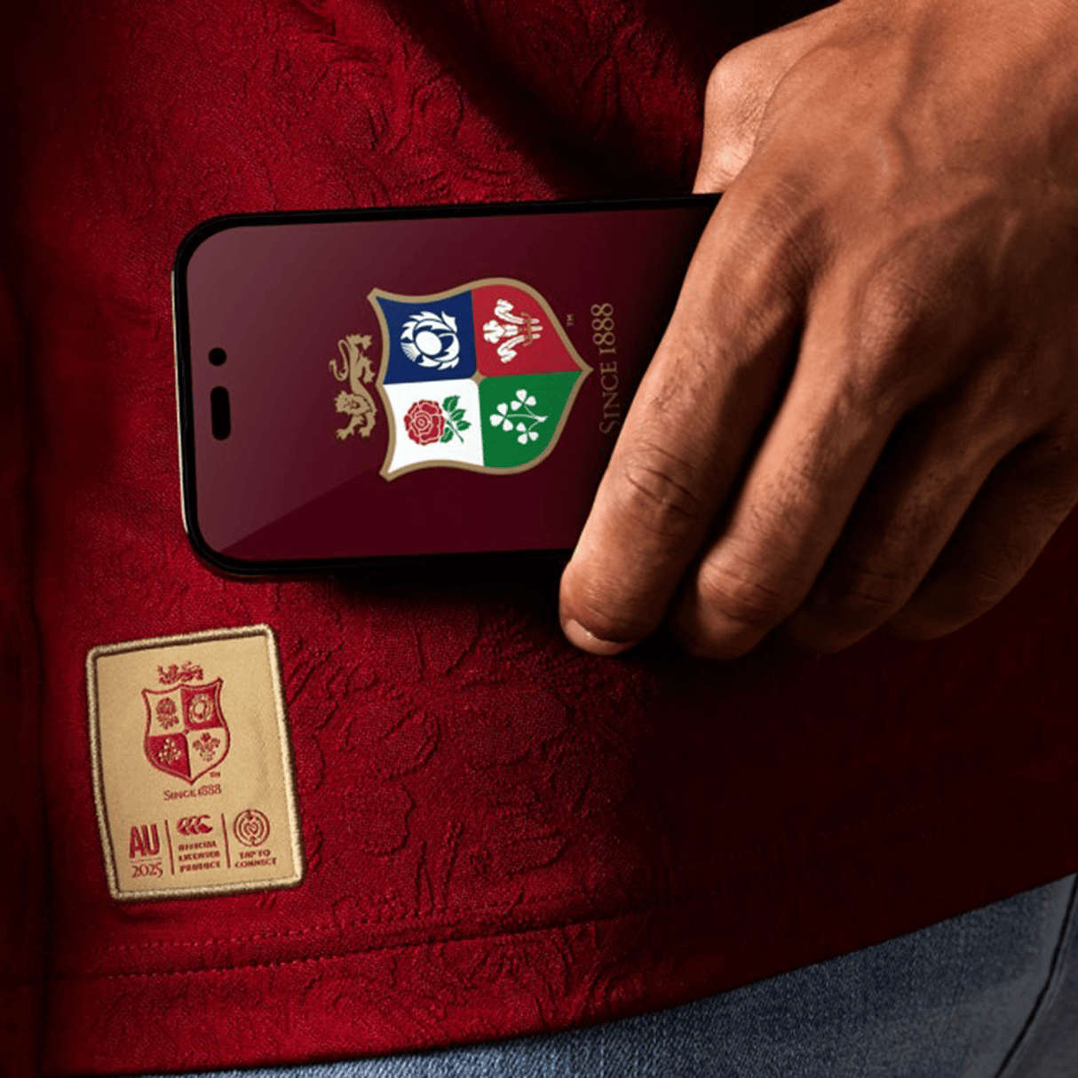 Close-up of a person holding a phone with a rugby-themed screen, wearing the red British & Irish Lions 2025 Players Jersey by Canterbury, featuring a stitched emblem.