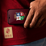 Close-up of a person holding a phone with a rugby-themed screen, wearing the red British & Irish Lions 2025 Players Jersey by Canterbury, featuring a stitched emblem.