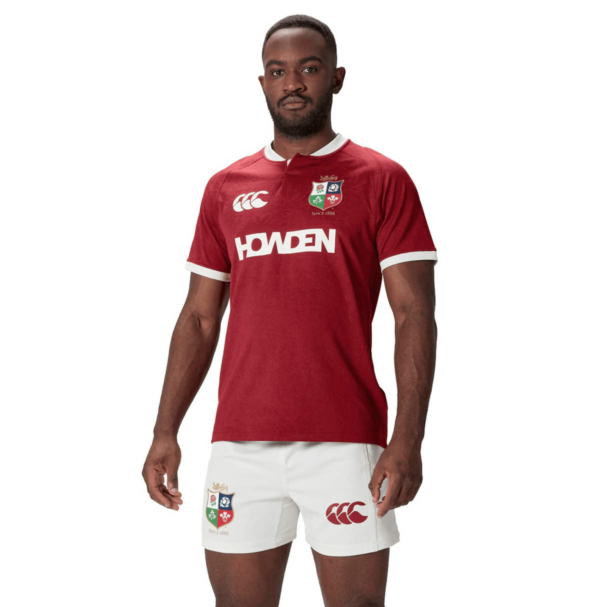 A person is wearing the British & Irish Lions 2025 Players Jersey by Canterbury, featuring a moisture-wicking red design with white trim and matching shorts, standing against a white background.