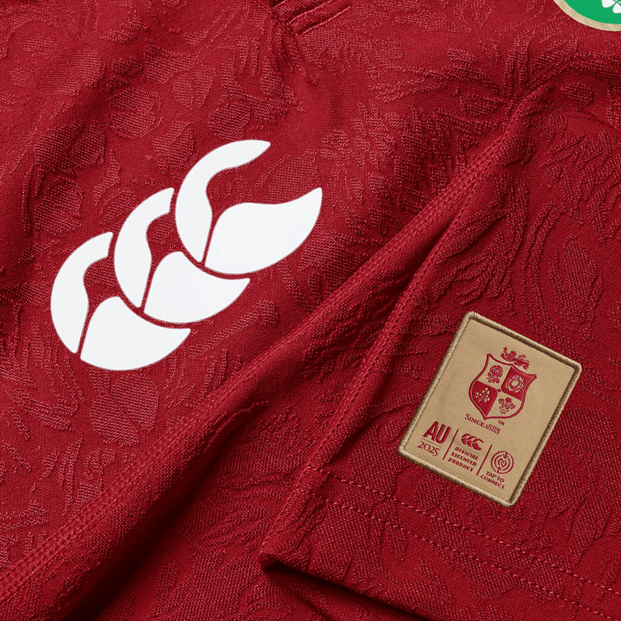 A close-up view of the British & Irish Lions 2025 Players Jersey by Canterbury, showcasing a red replica sports jersey made from moisture-wicking fabric. The design features a white logo with three curved shapes and a gold patch that displays a lion, reminiscent of the British & Irish Lions emblem, along with the text "AU 2025 Success.