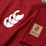 A close-up view of the British & Irish Lions 2025 Players Jersey by Canterbury, showcasing a red replica sports jersey made from moisture-wicking fabric. The design features a white logo with three curved shapes and a gold patch that displays a lion, reminiscent of the British & Irish Lions emblem, along with the text "AU 2025 Success.