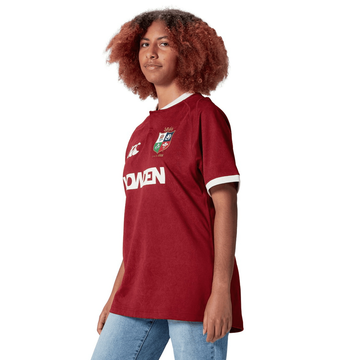 A person wearing a red British & Irish Lions 2025 Players Jersey by Canterbury and blue jeans stands against a white background.