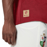 Close-up of a person wearing the British & Irish Lions 2025 Players Jersey by Canterbury, a red sports shirt crafted from moisture-wicking fabric featuring a gold label, paired with white shorts adorned with a colorful emblem.