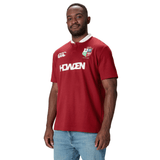 A man wearing the British & Irish Lions 2025 Classic Jersey by Canterbury stands against a white background, with its Red Dahlia tone adding a vibrant touch to his attire.