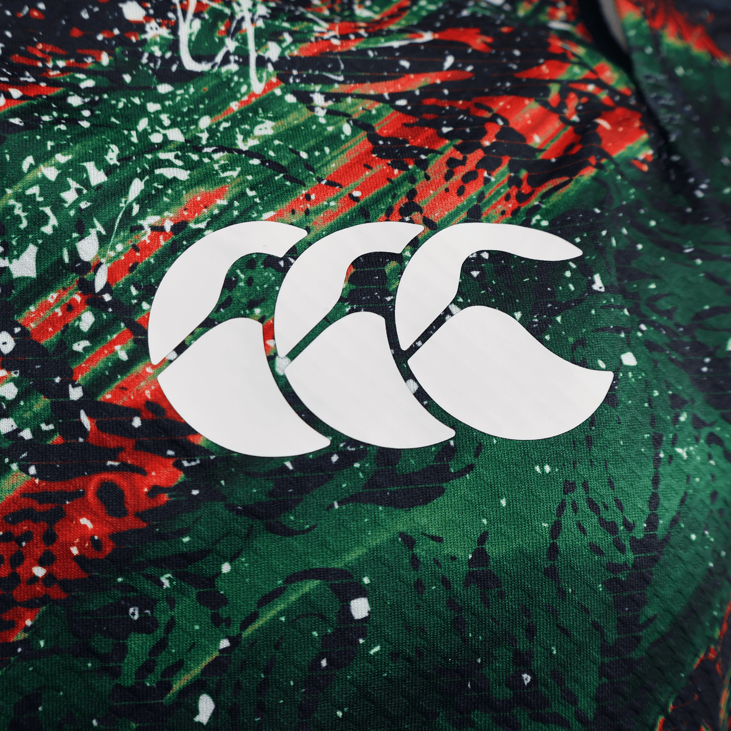 Close-up of the British & Irish Lions 2025 Replica Warm Up Jersey by Canterbury, featuring its red, green, and black pattern. The athletic fit fabric showcases a white logo with three curved shapes, designed for optimal performance with moisture-wicking properties.